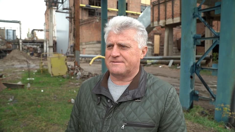 Mann mit weissen Haaren steht vor Metallstelzen und einem Gebäude aus Ziegelstein.