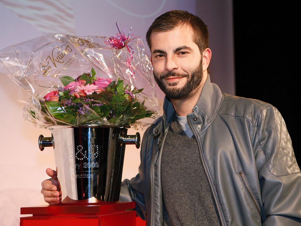 Bligg halbnahe in grauer Lederjacke, neben ihm auf dem Tisch steht der Glory-Pokal mit Blumenstrauss darin
