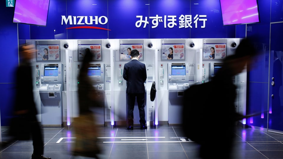 Fünf Geldautomaten einer japanischen Bank, ein Mann bezieht Geld, drei Personen gehen vorbei.