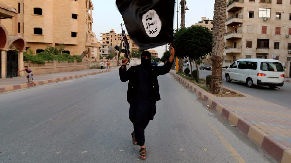 Mann in schwarz mit schwarzer IS-Flagge in einer Strasse, er ist bewaffnet.
