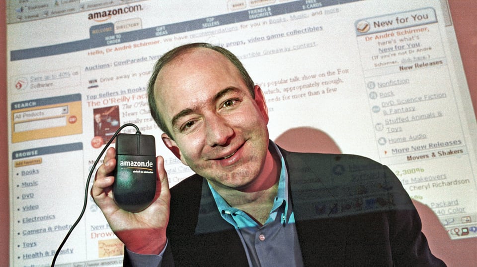 Mann mit wenig Haar, hält Computermaus mit Amazonlogo, trägt Anzug und hellbalues Hemd, dahinter Website-Screen.