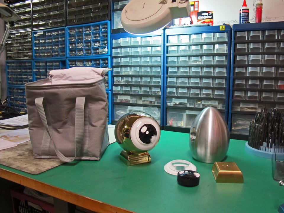 Ein Tisch, darauf v.l.n.r.: eine Kühltasche, ein Goldenes Auge, die Einzelteile des Auges (Aluminium-Ei, Sockel, Iris, Pupille)