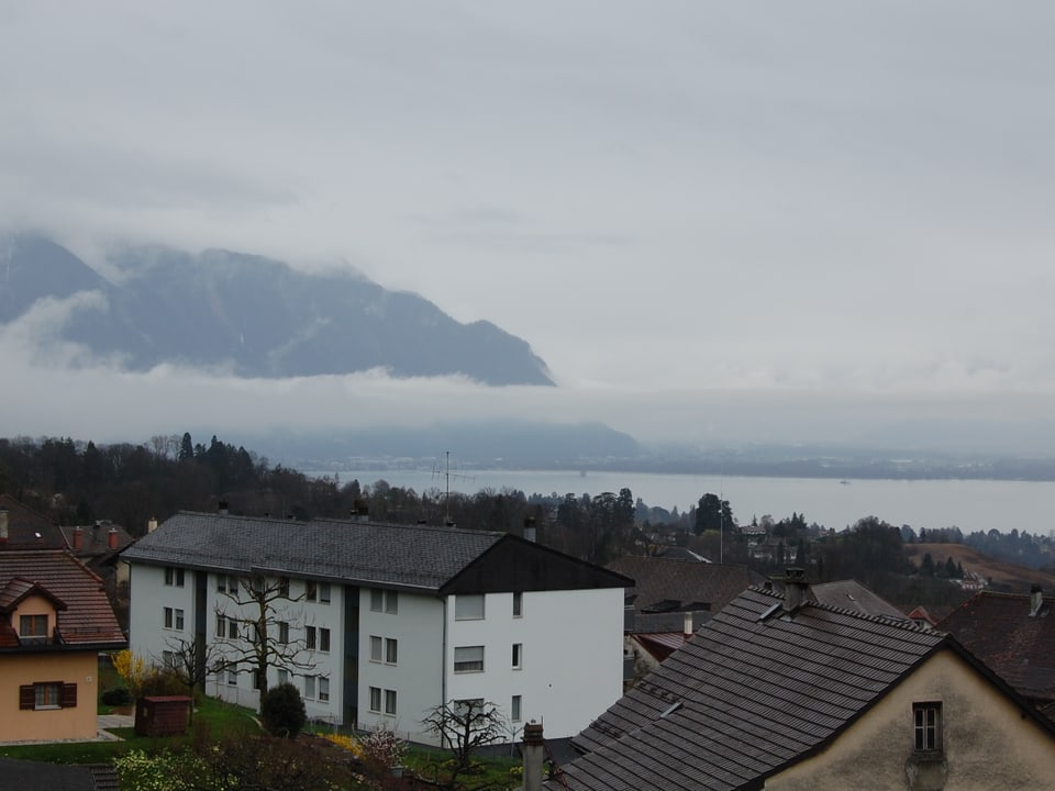 Hinter Häusern ist der graua Genfersee und in Wolken gehüllt einige Berge zu sehen.