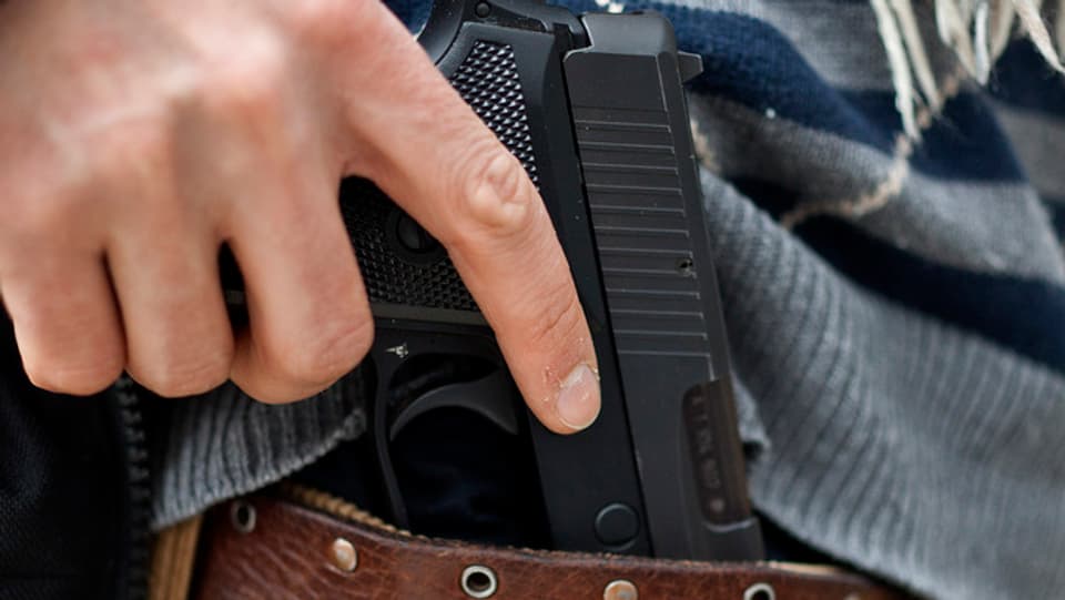 Eine Hand greift an eine Pistole, die in einem Hosenbund steckt. 
