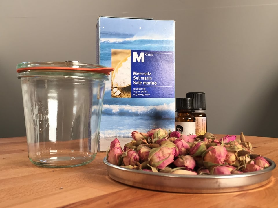 Meersalz, Rosenblüten, ein leeres Glas und ätherische Öle stehen auf einem Tisch.