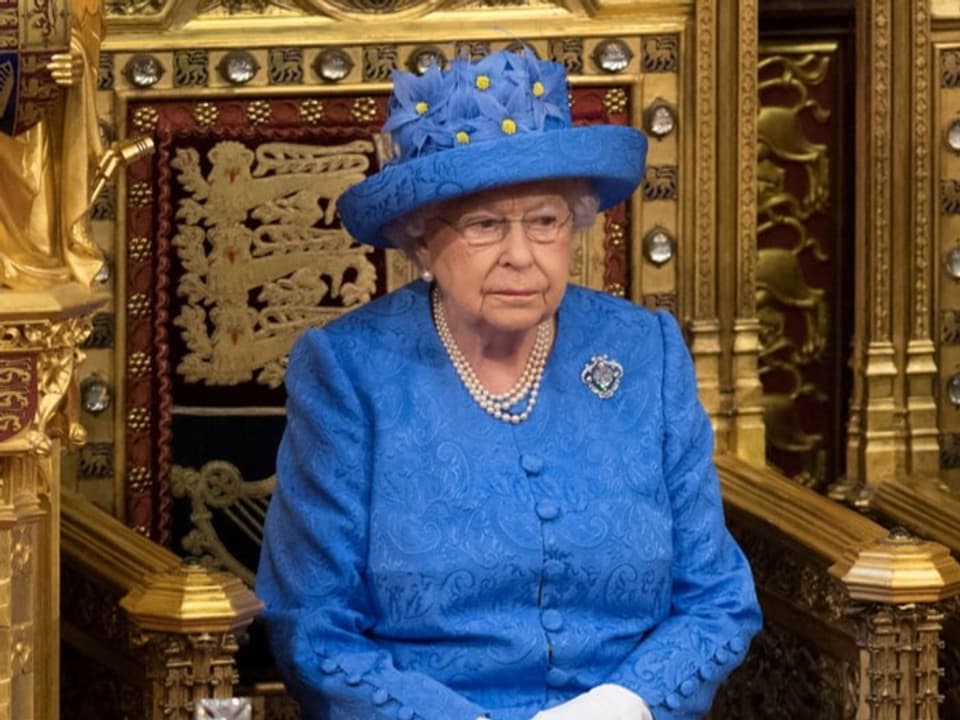 Queen Elizabeth in einem blauen Kleid und blauem Hut mit gelben Blumen.