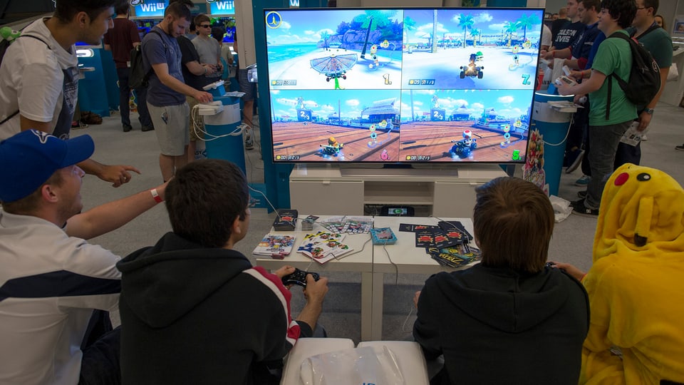 Vier Personen sitzen vor einem Bildschirm und spielen Mario Kart