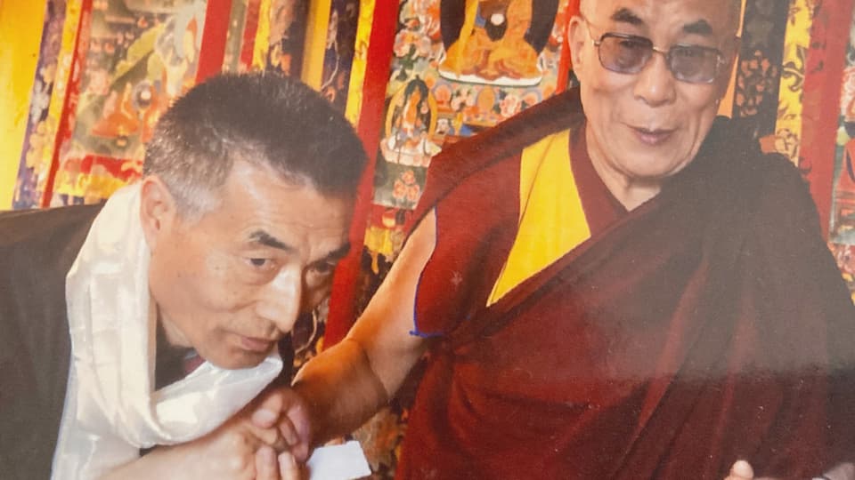Zwei Männer, der rechts trägt ein tibetisches Gewand und eine Sonnenbrille, der andere Mann schüttelt ihm die Hand
