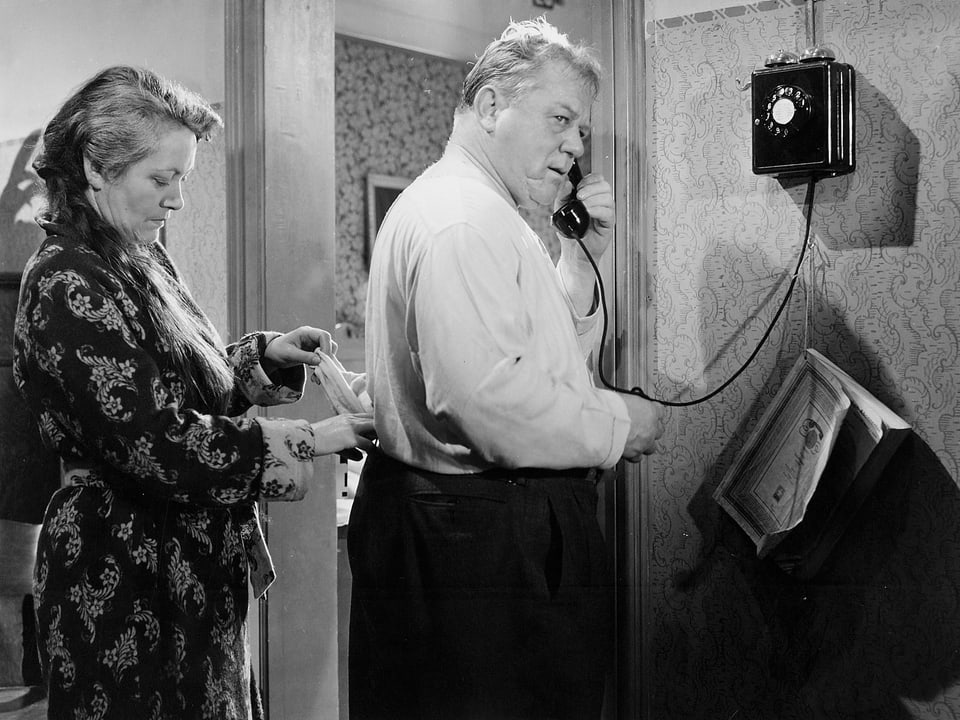 Schwarz-weiss Foto mit einer Filmszene. Schaggi Streuli am Wandtelefon, Margrit Rainer rückt ihrem Ehemann das Hemd zurecht.
