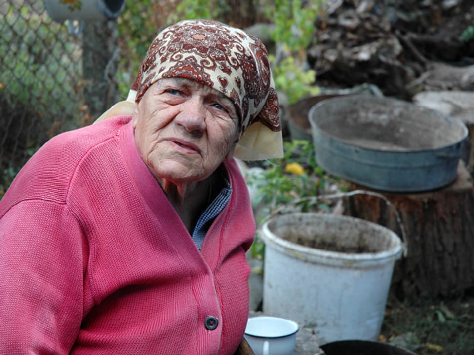 Eine alte Frau mit Kopftuch sitzt draussen im Garten.