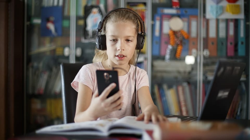 Ein Mädchen mit Kopfhörern und einem Smartphone sitzt vor einem Schulbuch.