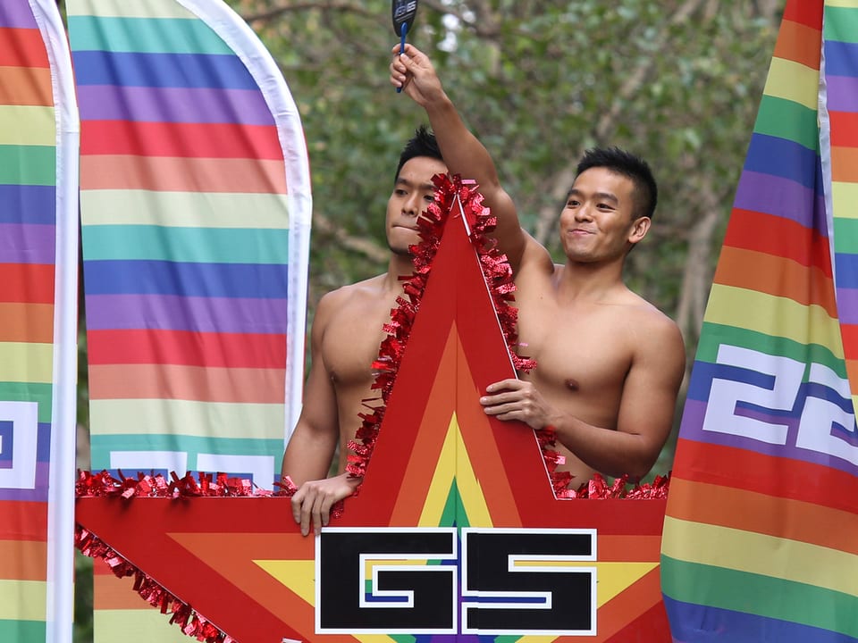 Zwei junge Männer mit nacktem Oberkörper demonstrieren zwischen regenbogenfarbenen Fahne.