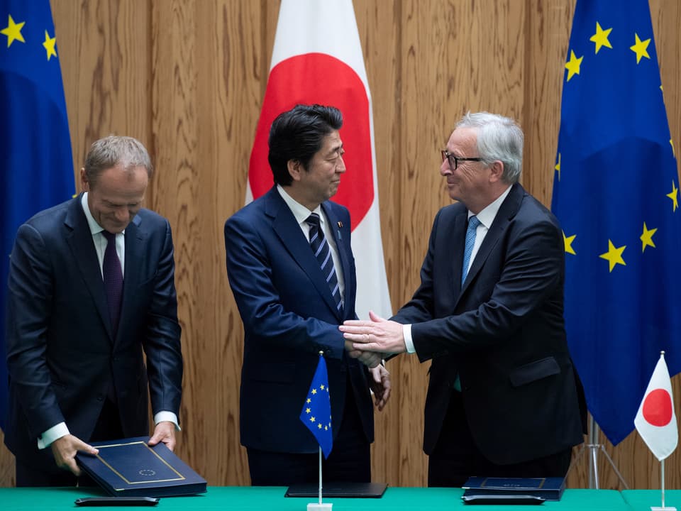 Tust, Shinzo Abe und Juncker stehen hinter dem Tisch. Juncker gibt Shinzo Abe die Hand.