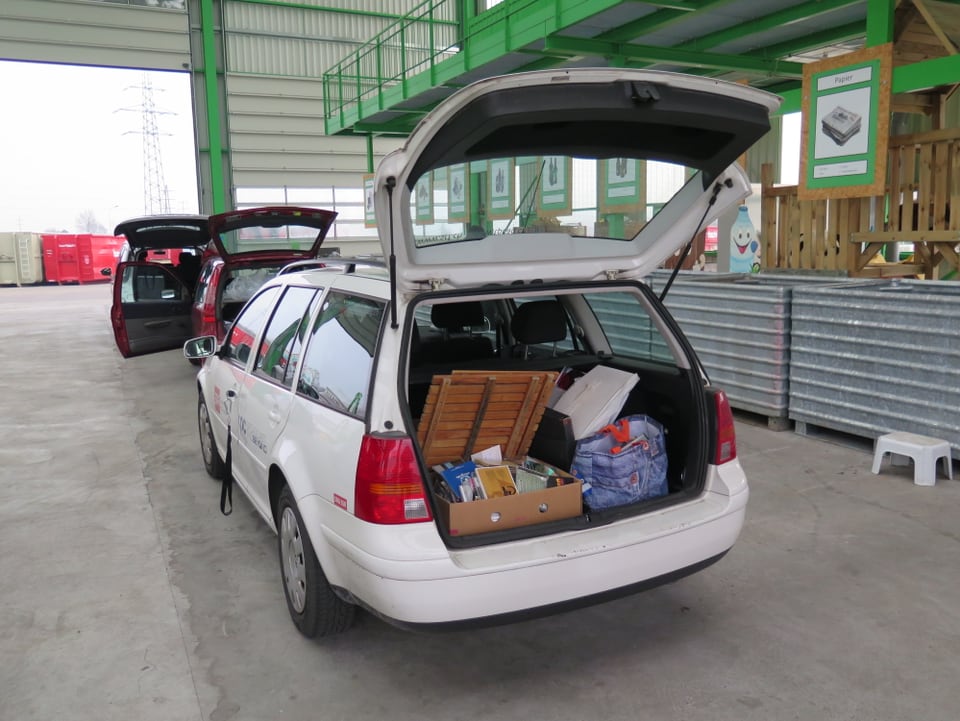 Weisses Auto mit geöffnetem Kofferraum, darin Holz, CD's und anderes Recycling-Material.