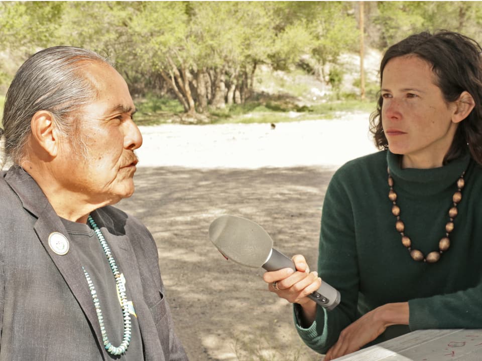 Frau Interviewt einen indianischstämmigen Mann, Bäume im Hintergrund.