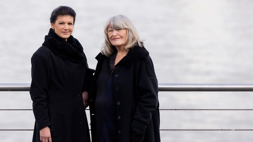 Alice Schwarzer steht neben Sarah Wagenknecht. Beide tragen einen schwarzen Wintermantel. Im Hintergrund der Rhein.