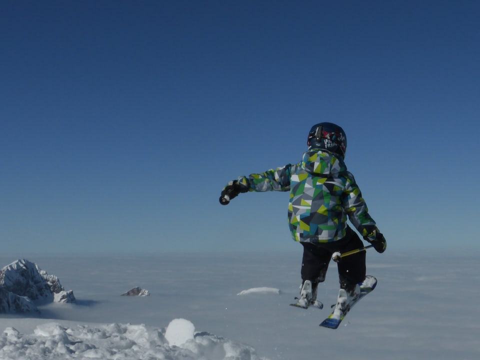 Wolkenloser Himmel, im Vordergrund ein Kind, das mit Skiern vom Berg herunter zu springen scheint. Tief unten ist das Nebelmeer zu sehen, heraus ragen ein paar Berggipfel. 