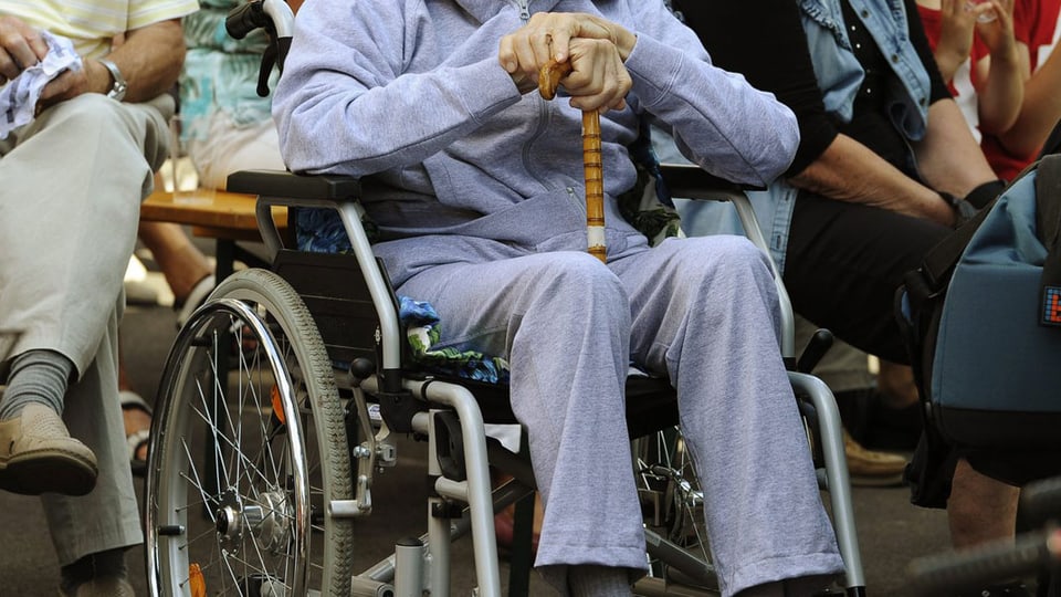 Ein alter Mann sitzt im Rollstuhl und stützt seine Hände auf einen Gehstock