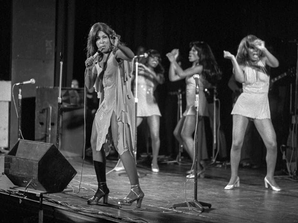 Schwarzweiss-Aufnahme von Tina Turner auf der Bühne zusammen mit Tänzerinnen.