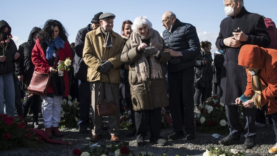 Die KZ-Überlebende Éva Pusztai-Fahidi inmitten von Menschen, vor ihren Füssen liegen Blumen.