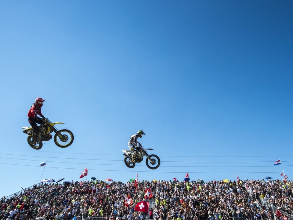 Zwei Motocrosser springen vor der Zuschauertribüne durch die Luft