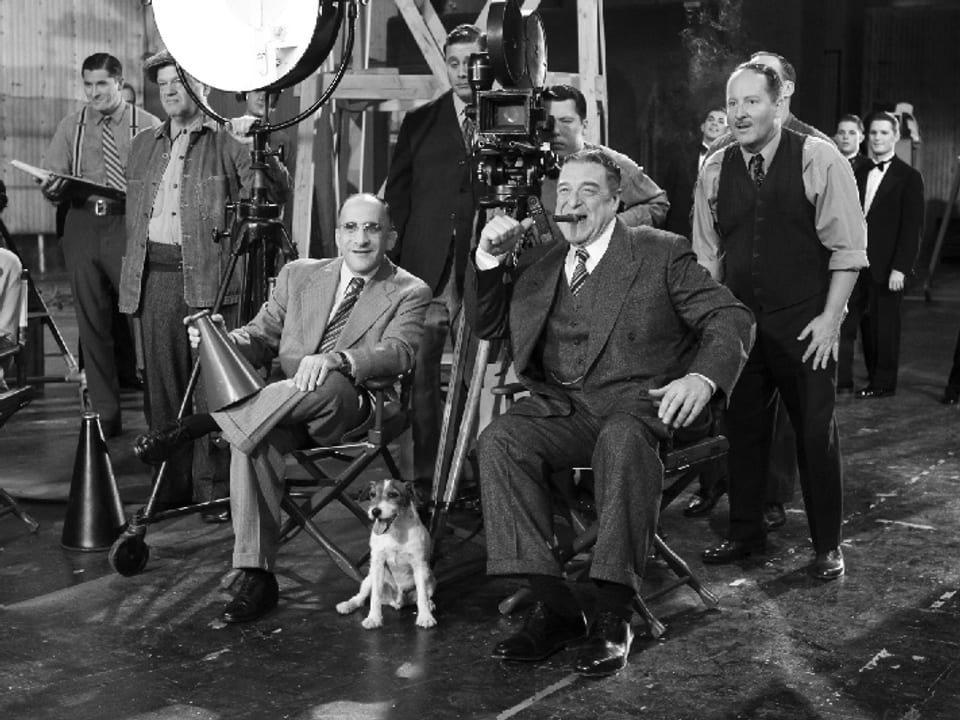 Szenen eines Drehs in Hollywood mit Kameramann, Regisseur und Assistenten. Ein zwei Männer sitzen in Vordergrund. Einer mit Zigarre im Mund, einer mit Megafon in der Hand. zu ihren Füssen sitzt ein kleiner, heller Hund.