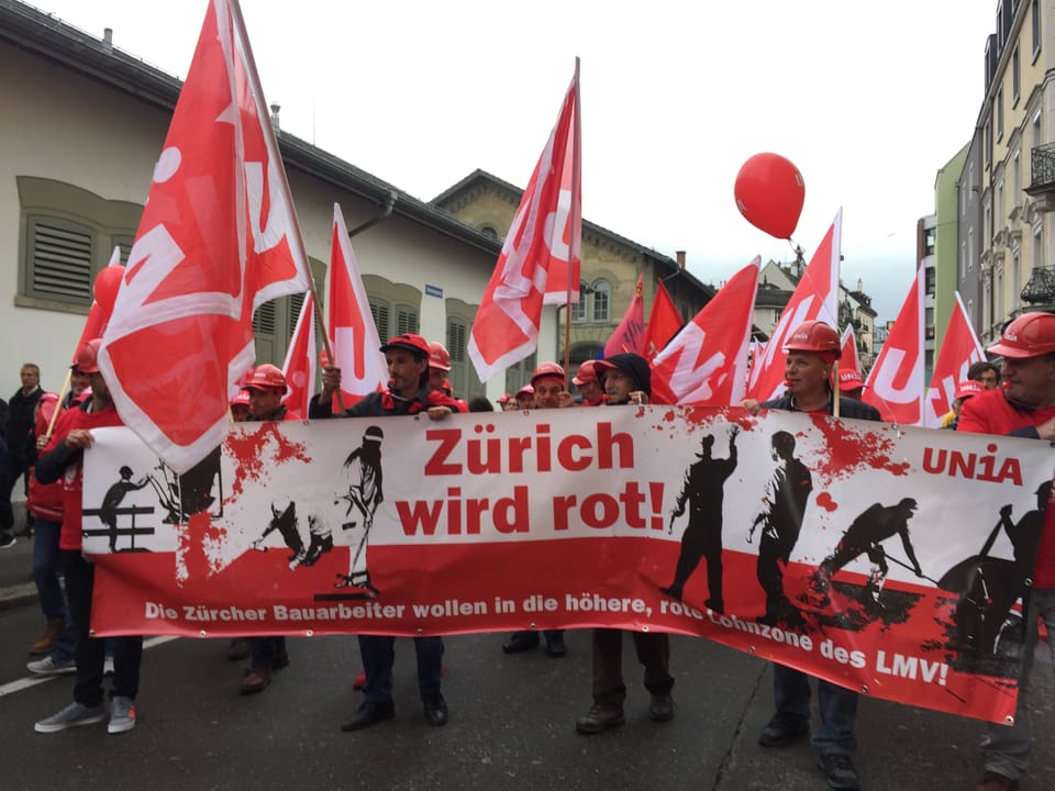 Männer mit Schutzhelmen und Unia-Fahnen halten ein Transparent mit dem Slogan "Zürich wird rot".