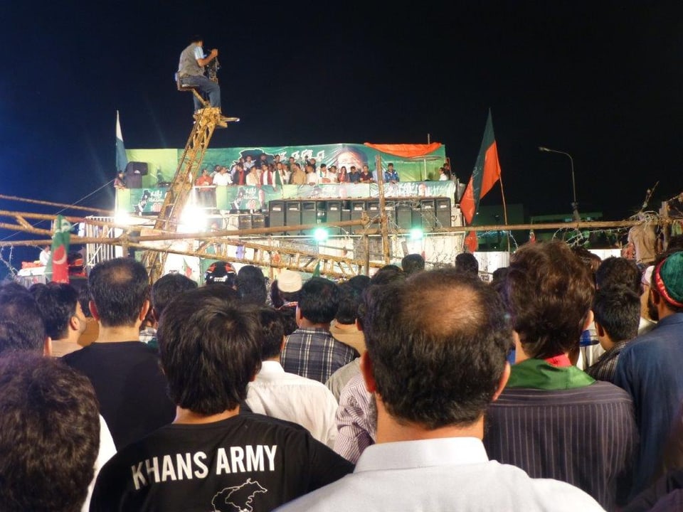 Menschen Blicken zu einem beleuchteten Klotz, auf dem eine Gruppe Leute steht, in der Mitte Imran Khan.