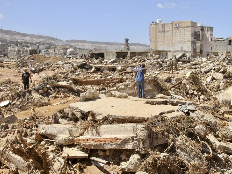 Die zerstörte Hafenstadt Derna. Einzelne Personen suchen in den Trümmern nach Überlebenden. In der Mitte des Bildes umarmen sich zwei Personen.