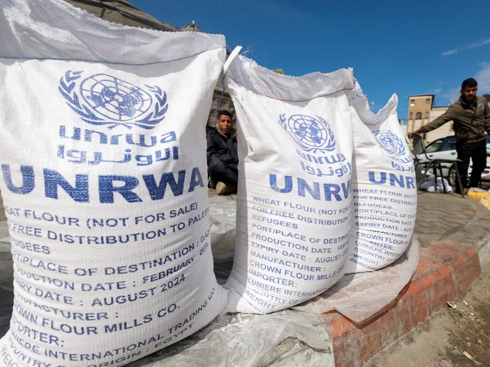 Aufnahme von Säcken mit Mehl. Sie sind mit UNRWA beschriften. Daneben stehen und sitzen Männer.