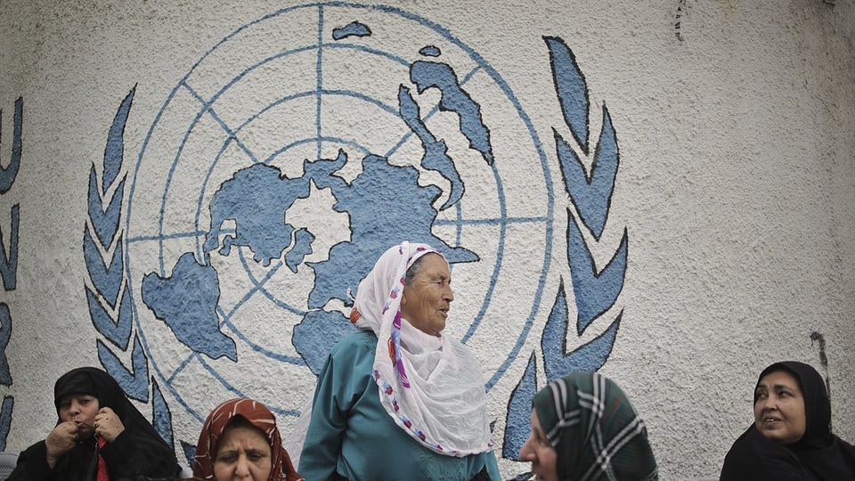 5 Frauen sind vor einer Wand, die mit dem UNRWA-Logo bemalen ist. Eine der Frauen steht höher in der Mitte.