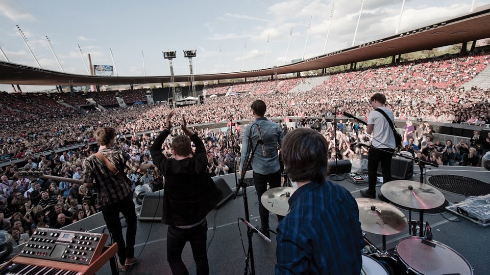 Blick von hinten auf eine Rockband, die in einem vollen Fussballstadion auf der Bühne steht.