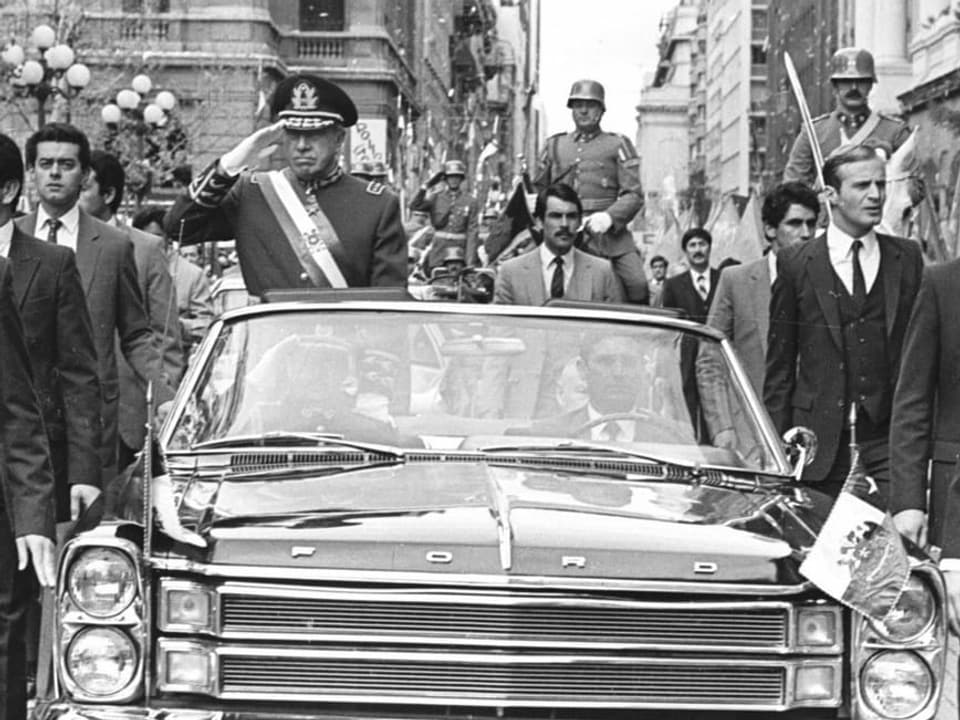 Bild in schwarz/weiss. Augusto Pinochet salutiert in einem fahrenden Auto. Das Auto fährt im Schritttempo durch eine Menschenmenge in der Innenstadt und wird begleitet von weiteren Abgeordneten, die neben dem Auto herlaufen. 