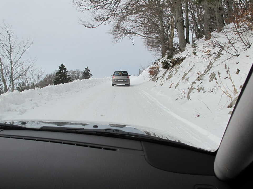 Foto aus dem Innern eines Autos durch die Frontscheibe nach draussen: Auto fährt auf schneebedeckter Strasse den Berg hoch. 