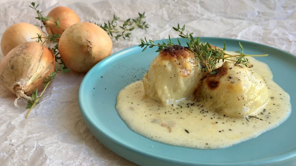 Ein graublauer Teller mit drei gratinierten Zwiebeln an einer Rahmsauce. Im Hintergrund weisse Zwiebeln und Thymian.