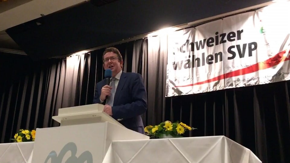 Ein Mann steht an einem Rednerpult und spricht in ein Mikrofon. Im Hintergrund das Plakat "Schweizer wählen SVP". 
