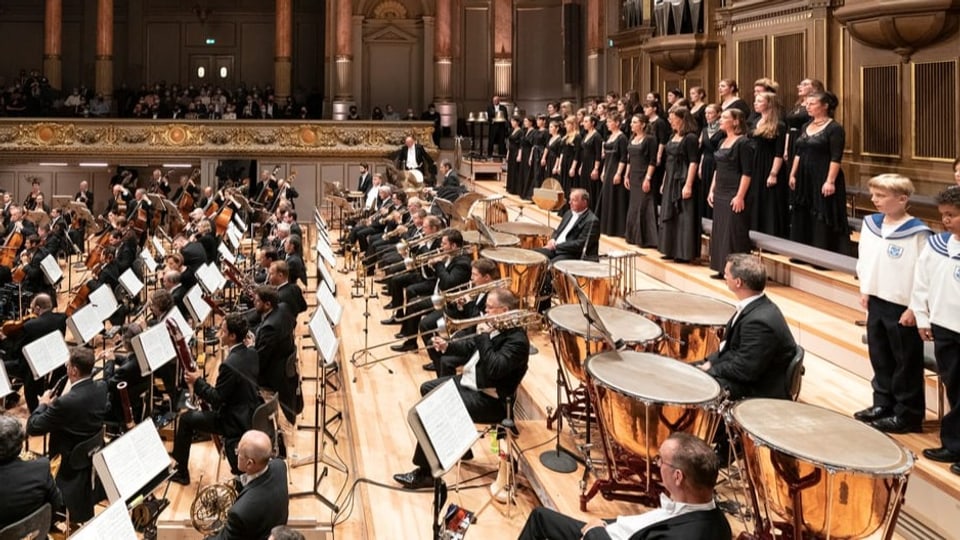 rechts stehende Frauen am Singen, links sitzende Musiker in Reihen und im Anzug.