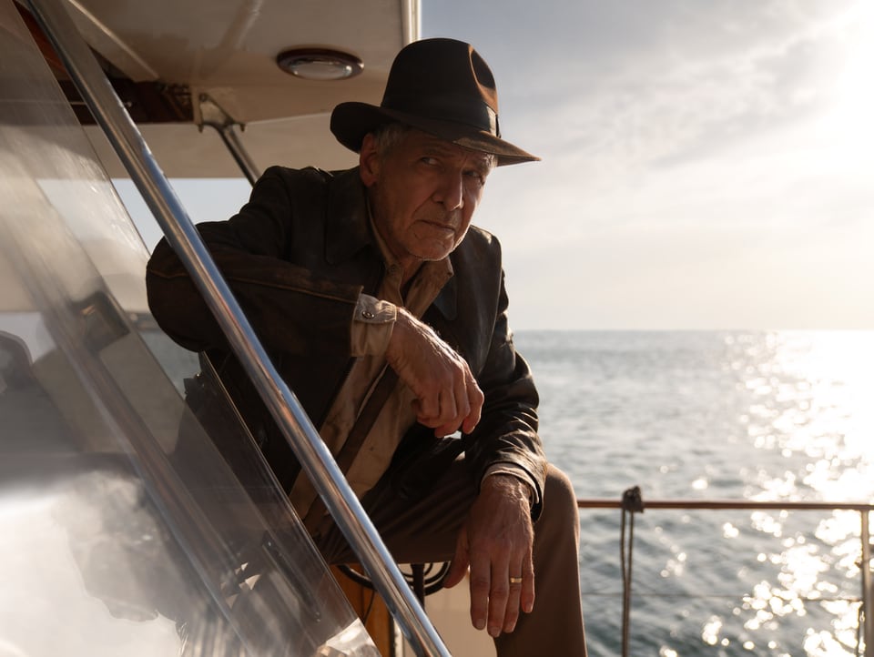 Grau melierter Mann mit Hut sitzt auf einem Boot in der Hocke und schaut skeptisch.