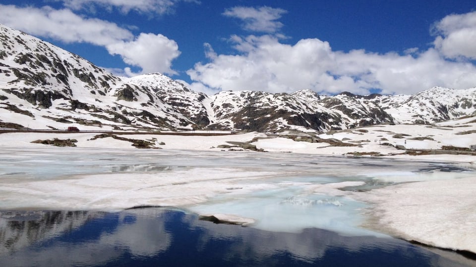 Schneeberge, wilde Wolken in blauem Himmel. Ein See, der zum Teil mit Eis bedeckt ist.
