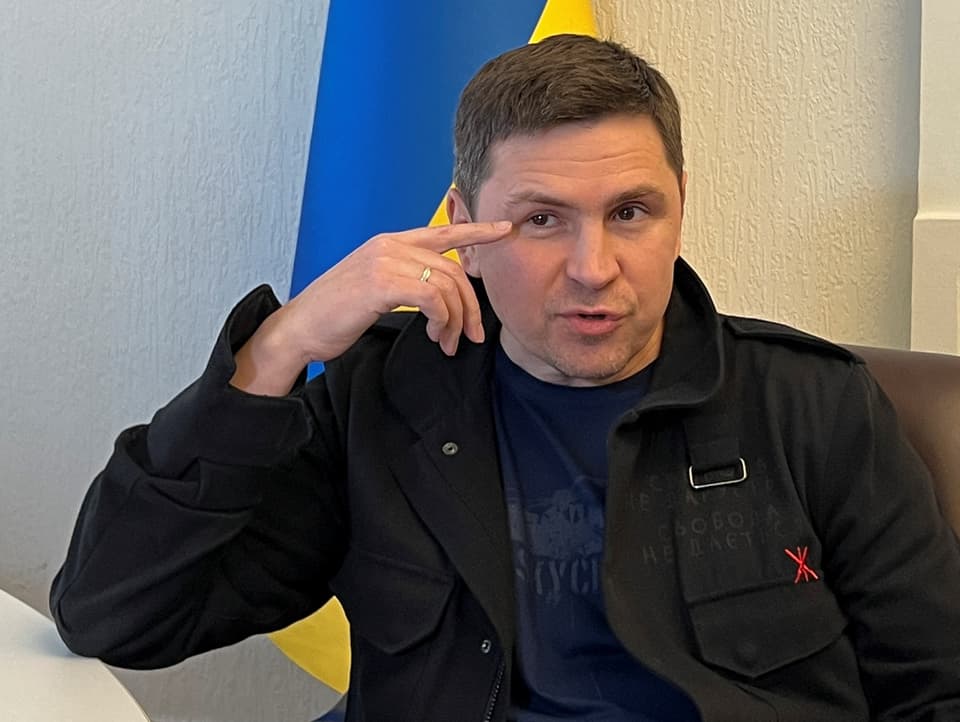 Mikhailo Podoliak im Bild, hinter ihm eine ukrainische Flagge. Dunkles Haar, der Zeitfinger streicht seine Augenlider.
