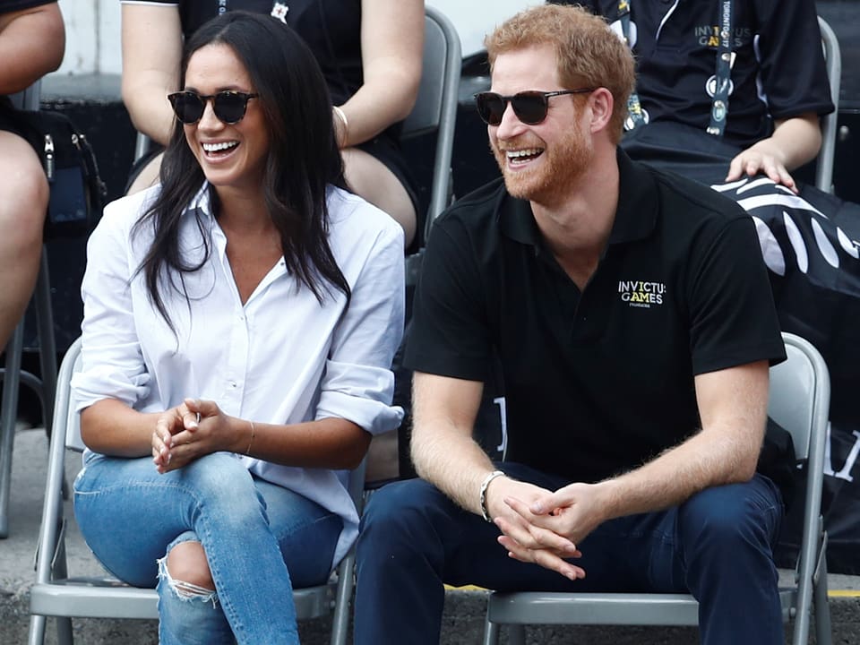 Meghan Markle und Prinz Harry mit Sonnenbrille auf Stühlen sitzend.