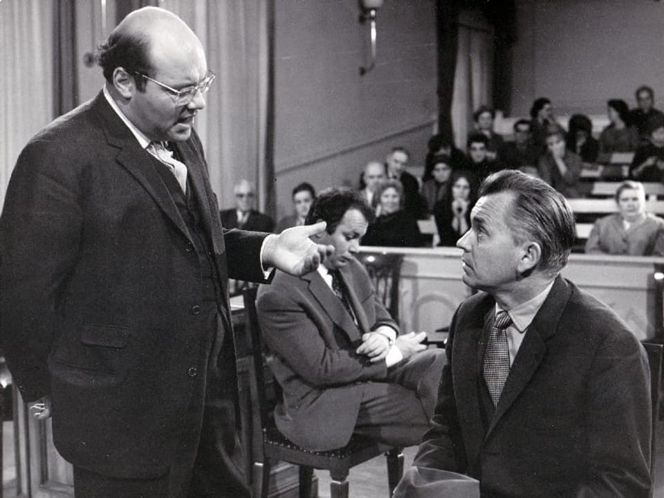 Ein glatzköpfiger Mann steht in einem Gerichtssaal und diskutiert mit einem sitzenden Mann. Im Hintergrund der Gerichtssaal.