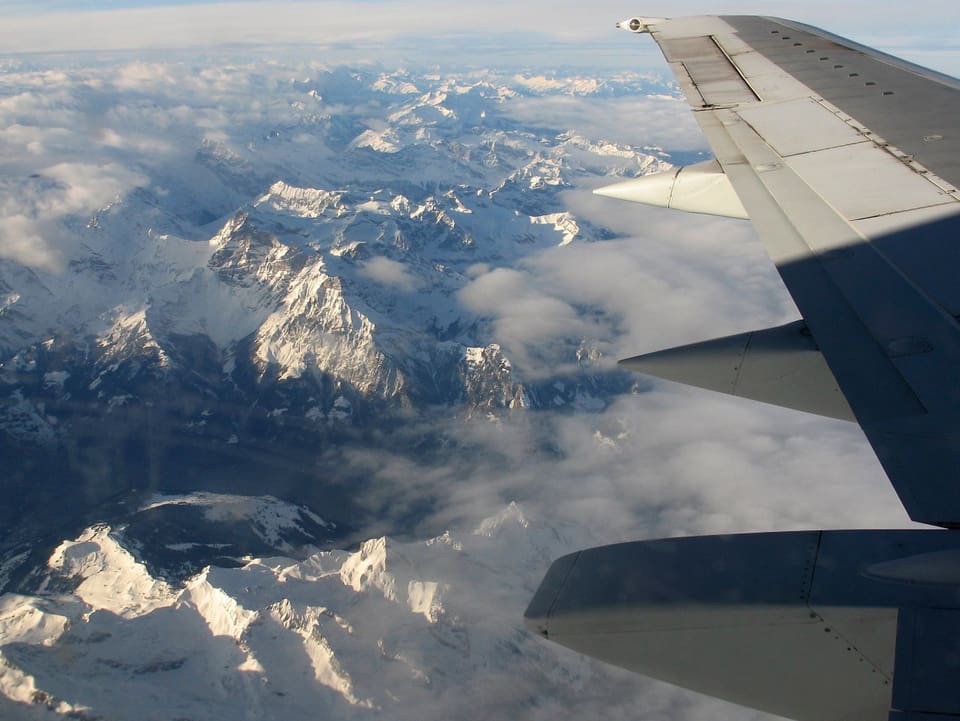 Blick aus dem Fenster eines Verkehrsflugzeuges auf die verschneiten Alpen aus grosser Höhe. Es hat auch einige Wolken. Rechts ist noch ein Teil des Flügels zu sehen. 