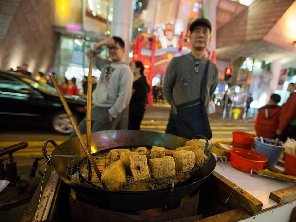Ein Mann steht hinter einem Wok, in dem rechteckige Stücke Tofu frittiert werden.