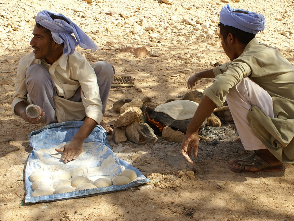 Zwei Beduinen sitzen ums Feuer, auf dem gerade ein Fladenbrot gebacken wird.