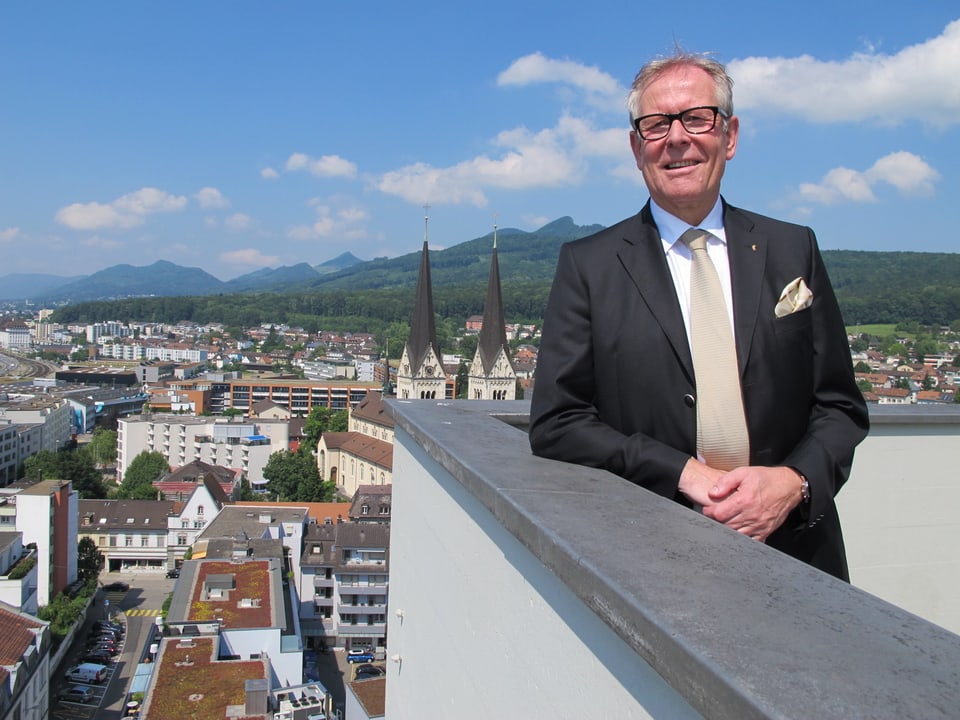 Ernst Zingg trägt einen Aanzug mit schwarzem Blazer, weissem Hemd, beiger Krawatte und beigem «Poschettli». Im Hintergrund die Martinskirche und die Stadt Olten, sowie die Jurakette.
