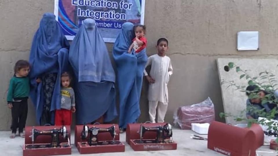 Frauen in Burkas mit ihren Kindern. Davor rote Nähmaschinen.