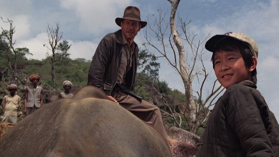 Indiana Jones sitzt auf einem Elefantenrücken, rechts von ihm ein kleiner Junge mit Baseballkappe