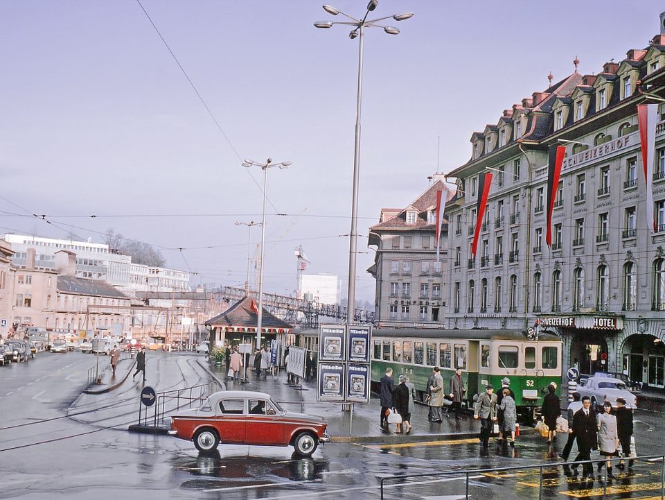 Grünes Zügli hält vor grossem Hotel, davor ein rotes Auto aus den 1960er Jahren. 