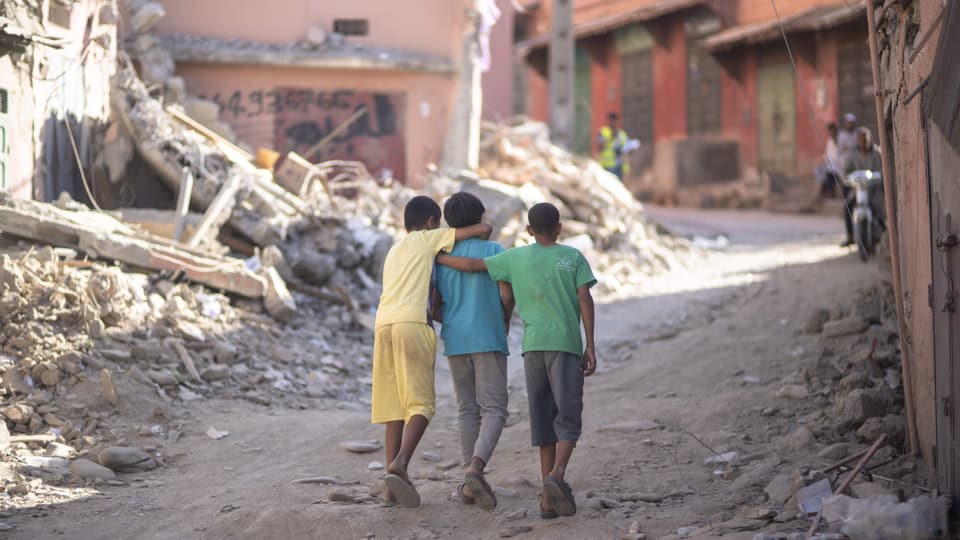 Drei Buben laufen Arm in Arm (von hinten fotgrafiert) durch Trümmerlandschaft in einer Ortschaft